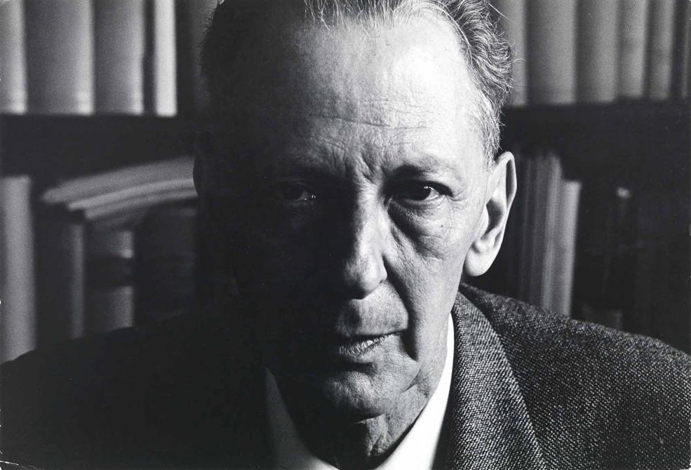 Black and white photograph: Portrait of a middle-aged man in front of a wall of books. One half of his face is heavily shaded.