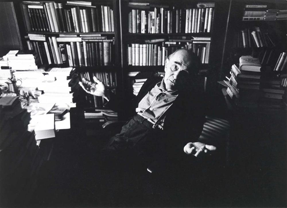 Black and white photograph: An elderly man sits in front of a wall of books in a study. Many books can also be seen on the table. He is photographed from diagonally above, holding both his arms out to the side with his palms open upwards.