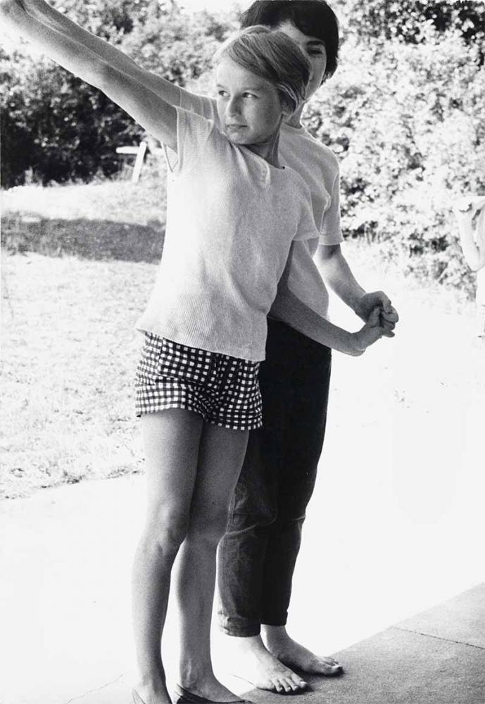 Black and white photograph: Two children stand behind each other. They are holding hands on the right and holding their arms up at an angle on the left. It looks as if they are performing something. Behind them, the sun is shining on a meadow and a hedge.