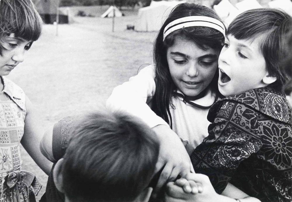 Black and white photograph: Four children at play, three of them standing close together, some with their arms crossed or holding hands. A girl stands to the left of the group and looks at the other knotted children. In the background, tents on a meadow can be seen in a blur.