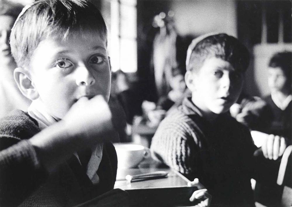 Black and white photograph: Several children are sitting at tables, but only the boy at the front left of the picture is in focus. He appears to be eating and puts his fingers in his mouth. He is wearing a kippah, as is the boy behind him on the right. The sun is shining on their faces from the left.