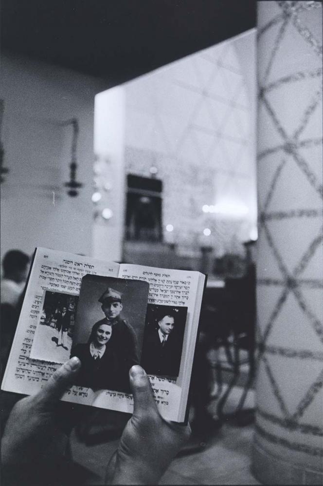 Black and white photograph: In the foreground of the picture, hands can be seen holding an open Hebrew prayer book up to the camera. Inside are three photographs: the first shows a man in a suit standing on a path, the second is a double portrait of a man in uniform and a woman in dark clothing with a light collar, the third is a portrait shot of a man in three-quarter profile with a dark suit, tie and shirt.