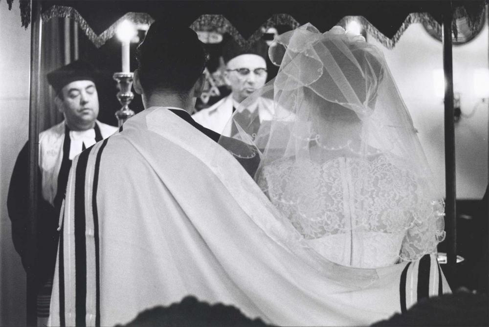 Black and white photograph: A bride and groom stand under a wedding canopy, both their backs facing the viewer and partially covered by a prayer shawl. The rabbi who is marrying them and a chasan (cantor) are standing in front of the bride and groom.