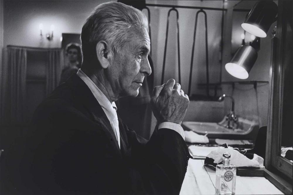 Black and white photograph: An older man sits at a table in a theater dressing room and stares into the mirror. He is seen in profile, wearing a jacket, his gray hair combed back. He is resting his elbows on the table, his hands are touching and appear slightly clenched.