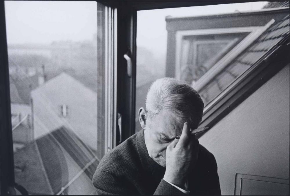 Black and white photograph: An older man with short gray hair wearing a jacket with a woven pattern is sitting in the dormer window of a house. He keeps his head down and touches his forehead thoughtfully. He is holding a cigar or a pen in his hand. The roof and neighboring houses can be seen through the window in the background.