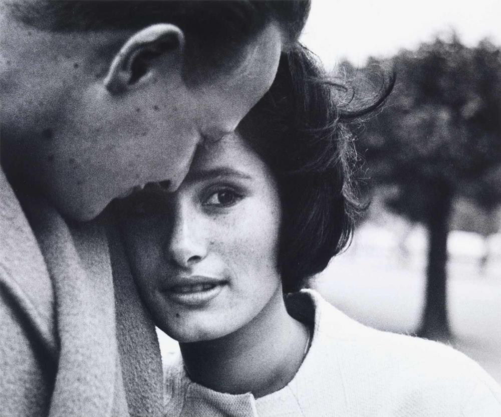 Black and white photograph: Portrait of a young couple: She leans her face against his shoulder and looks into the camera, his face is turned towards her and can be seen in profile from diagonally behind, his head is leaning against hers.