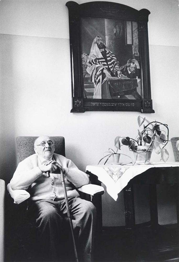 Black and white photograph: An old man is sitting in an armchair, holding a walking stick in front of him. To his right is a table with a tablecloth and two potted plants on it. A large painting hangs on the wall above him. It shows a scene from a synagogue. The painting is framed in a heavy frame with Hebrew lettering.