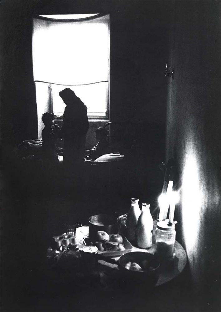 Black and white photograph: A woman is standing in front of a window in a darkened room. She is looking down at a child squatting on a bed in front of her. Another child is sitting to her right. In the foreground is a candlelit table. It is set with fruit, milk and other things.