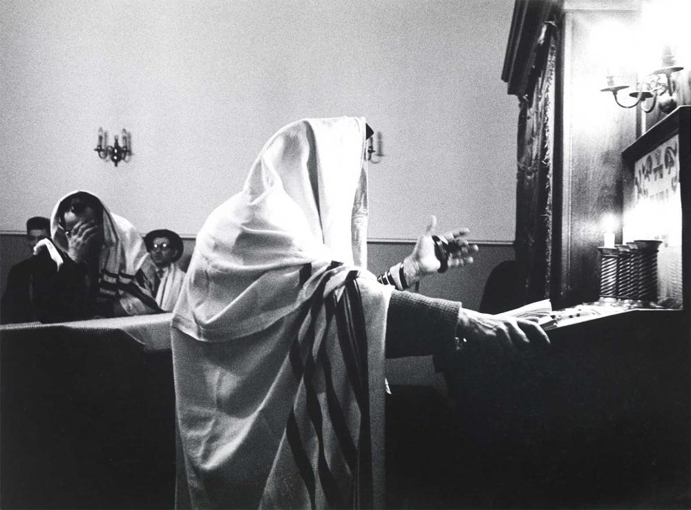 Black and white photograph: A prayer leader stands in front of the lectern in a prayer room. His head is covered with a tallit (prayer shawl) and his wrist is wrapped with a prayer strap. The lectern is lit by candles. Three men sitting on benches can be seen in the background. The man in front covers his face with his right hand.