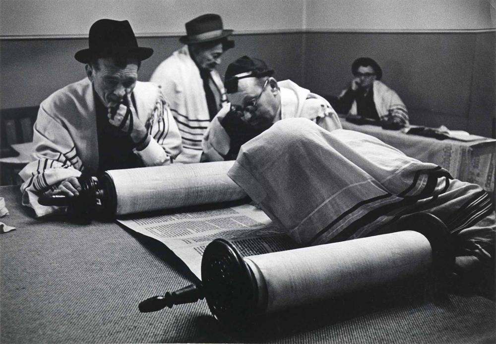 Black and white photograph: Five men are in a room. All are wearing headgear and a tallit. Four are sitting, one is standing. The face of the person in the foreground is completely covered by the tallit. He is reading a scroll.