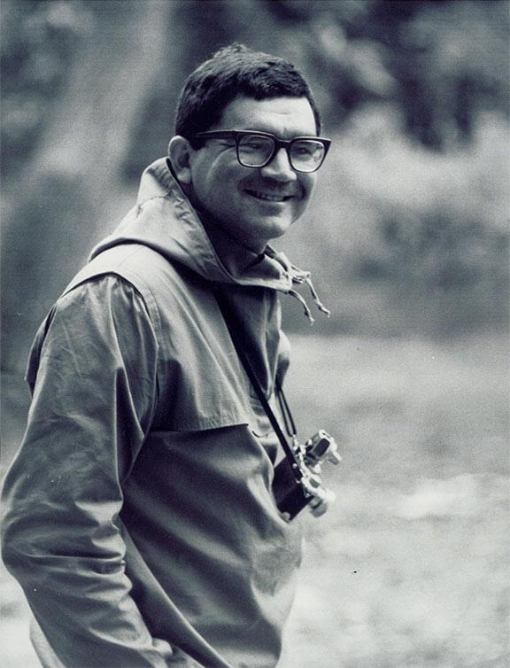 Black and white photograph of a man (Leonard Freed) looking into the camera and smiling. His upper body can be seen, he is wearing a jacket and glasses and has a camera around his neck. He is standing outdoors, the landscape in the background is blurred.