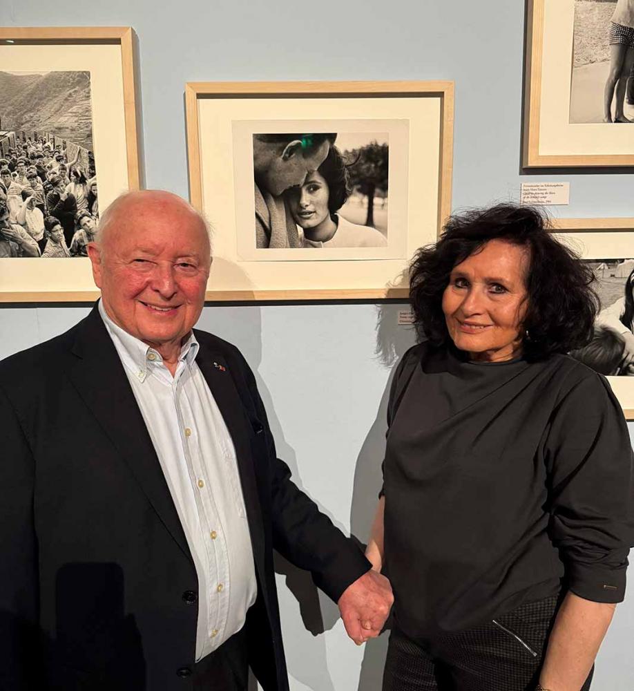 An older couple (man and woman) are standing in a photo exhibition holding hands in front of a black and white photo in which they are depicted as a young couple.