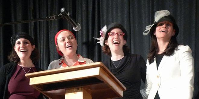 Four young women with hats or other headgear laughing behind a lectern with a microphone.