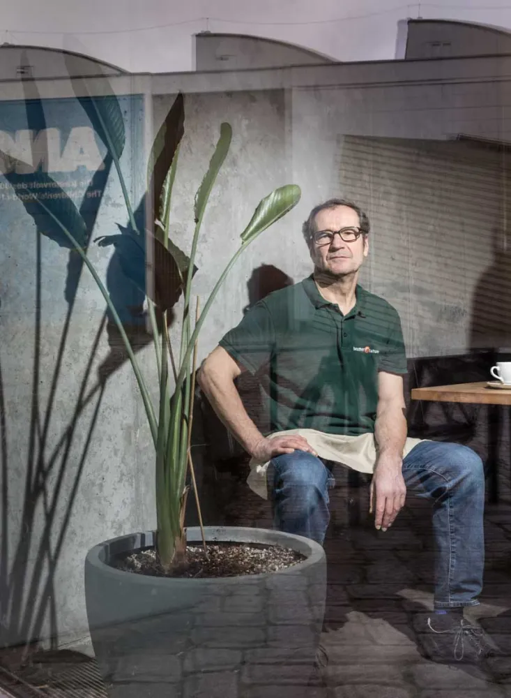 A man in a green shirt with the inscription Beumer&Lutum is sitting at a table. Next to him is a large houseplant.