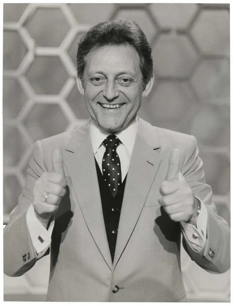 The black and white photo shows a half-portrait of a man in suit and tie with both thumbs up and looking friendly into the camera, with honeycomb pattern in the background.