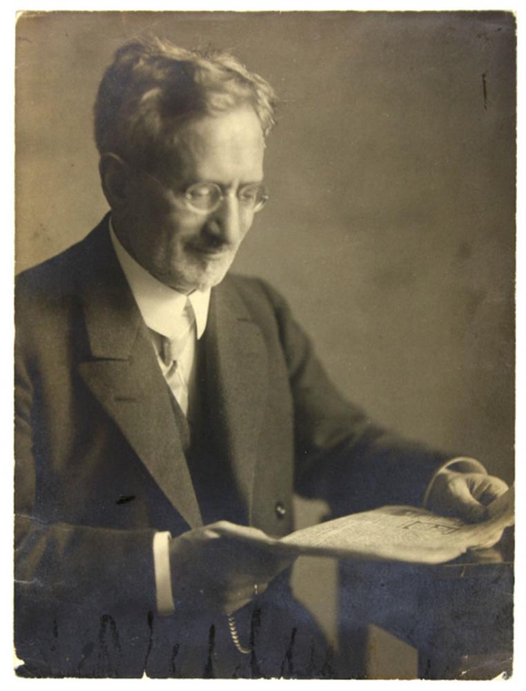 Black-and-white photograph of a man wearing a suit and glasses reading a newspaper while sitting at a table.