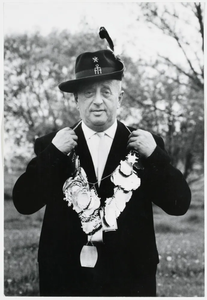 Black and white photograph: A gentleman in a hunting hat and suit presents a necklace with numerous pendants.
