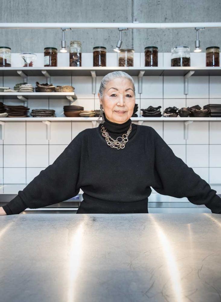 A woman with gray hair, a black top and a gold chain stands in a large kitchen behind a silver countertop.