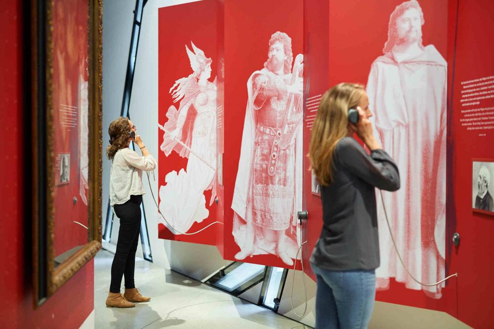 Two visitors stand with listeners in their hands in front of a red wall with white figures