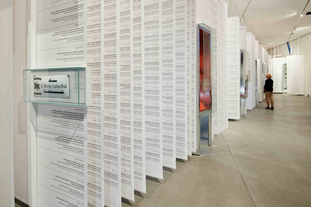 Long white paper banners with text hang from the ceiling interrupted by showcases, in the background stands a blonde woman