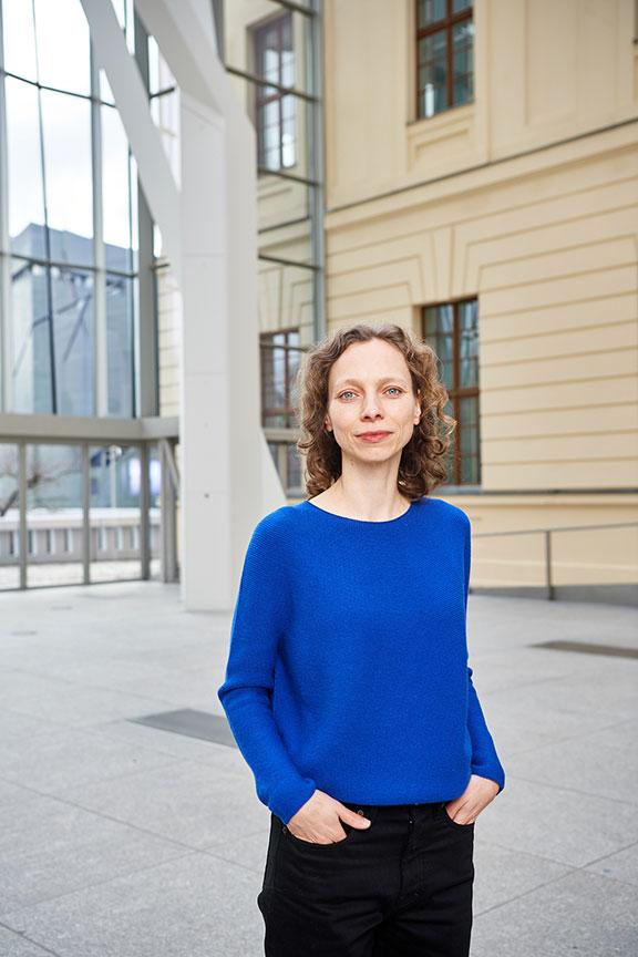 Portrait photo of a smiling woman wearing a bright blue jumper and black trousers: she looks directly into the camera, her hands are loosely in her trouser pockets. 