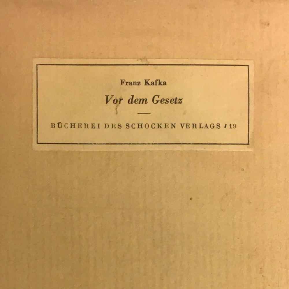 Detail of a yellowish-beige book cover with a stick-on label that reads: “Franz Kafka, Vor dem Gesetz (Before the Law), Bücherei des Schocken Verlags / 19th century.”