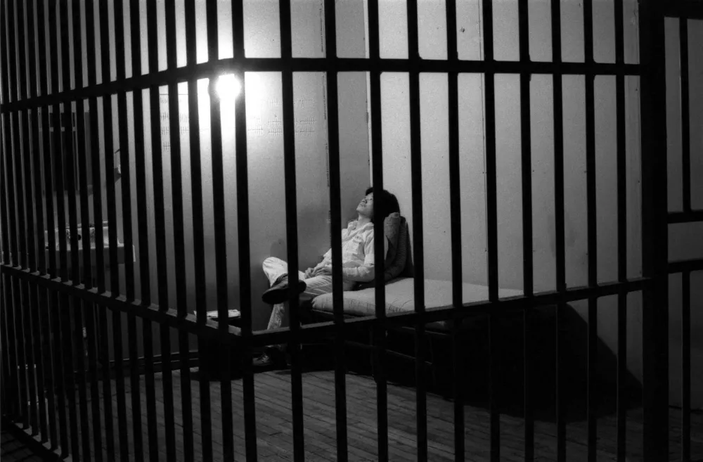 Black and white photo of a person behind bars, sitting on a cot.