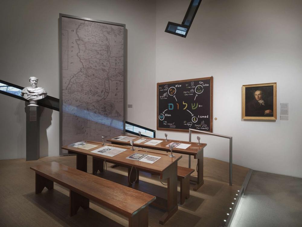 Wooden desks sit before a chalkboard filled with colorful Hebrew letters