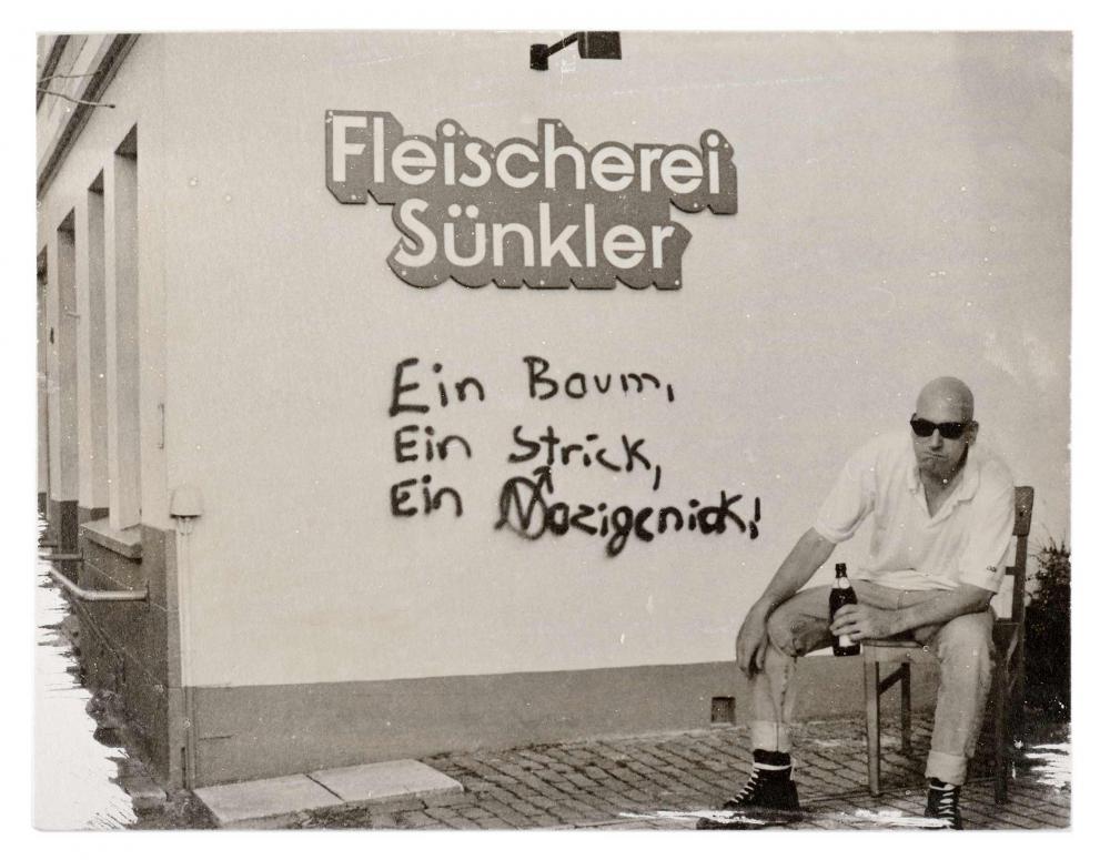 Ein Mann mit Sonnenbrille und einem Bier in der Hand sitzt neben einer Wand mit Graffiti.