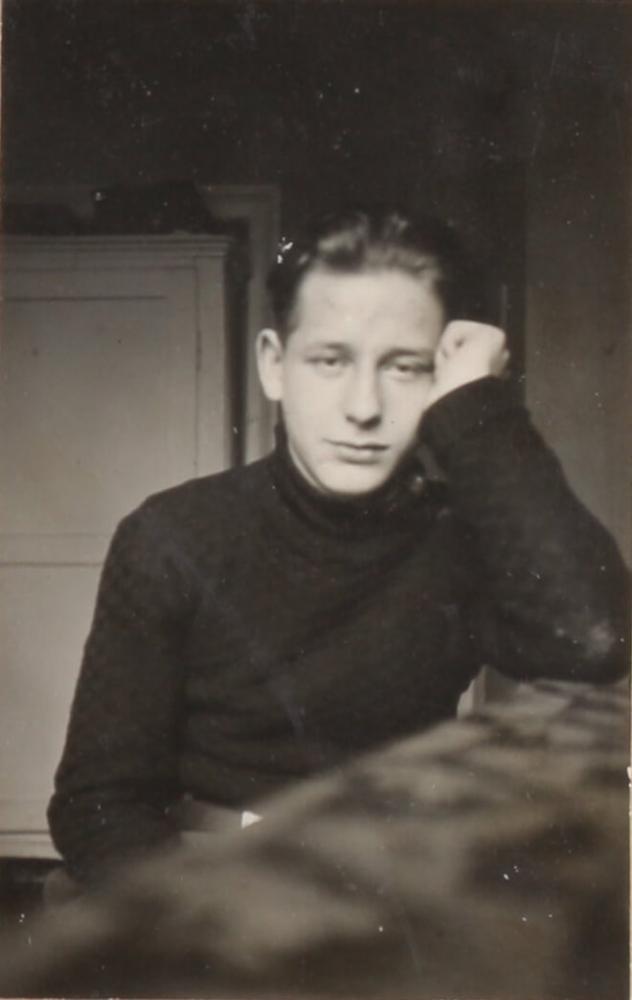 Black-and-white portrait photograph of a young man looking straight into the camera. His temple is propped up by the first of his left hand, his elbow resting on the table, where he is seated.