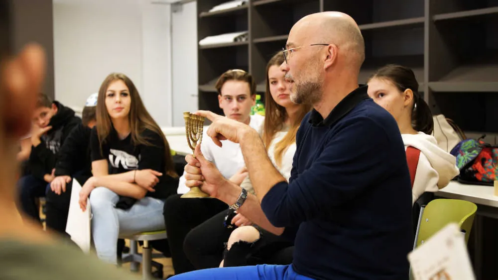 A man wearing glasses holds a Hanukkah candelabra in one hand while talking. A group of young people sits around him.