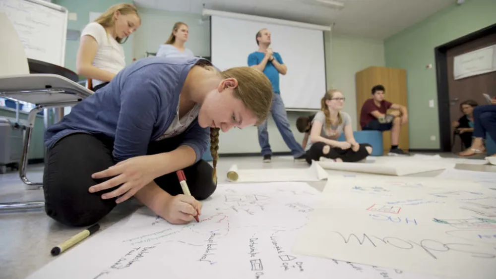 Young people and an adult are in a room with a projector screen and board. Posters with writing on them are spread across the floor. In the foreground, a girl sits on the floor writing on a poster.