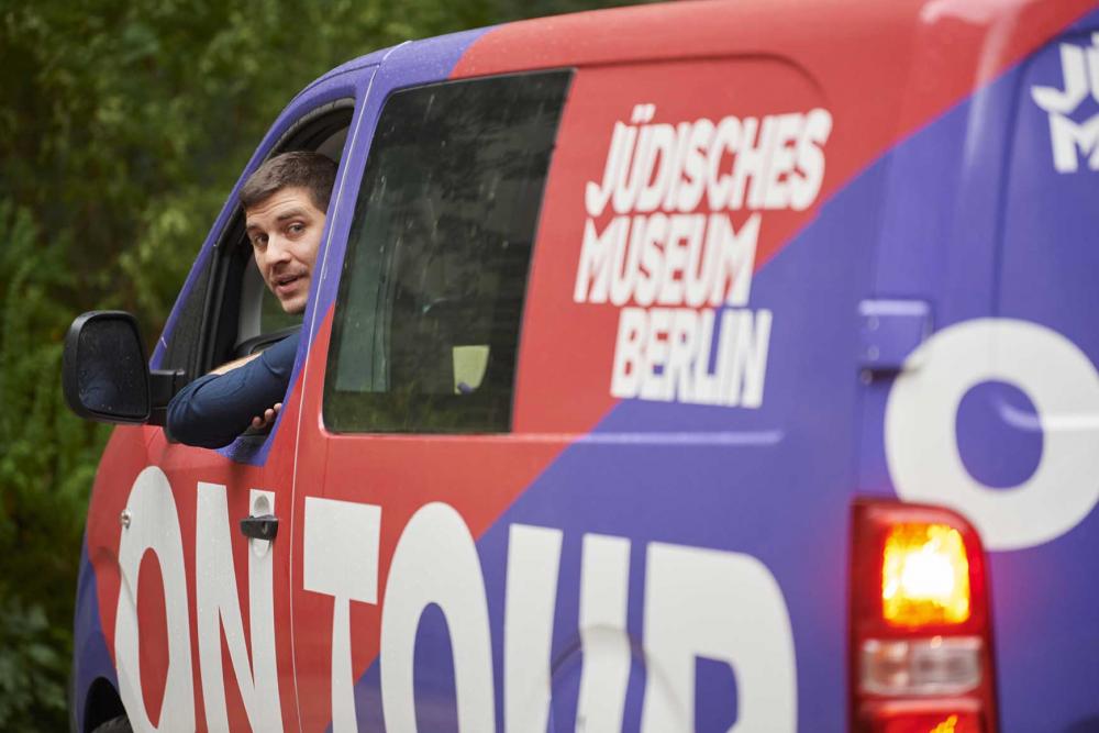 A man looks out of the window of a small van labeled “Jewish Museum Berlin on.tour”.