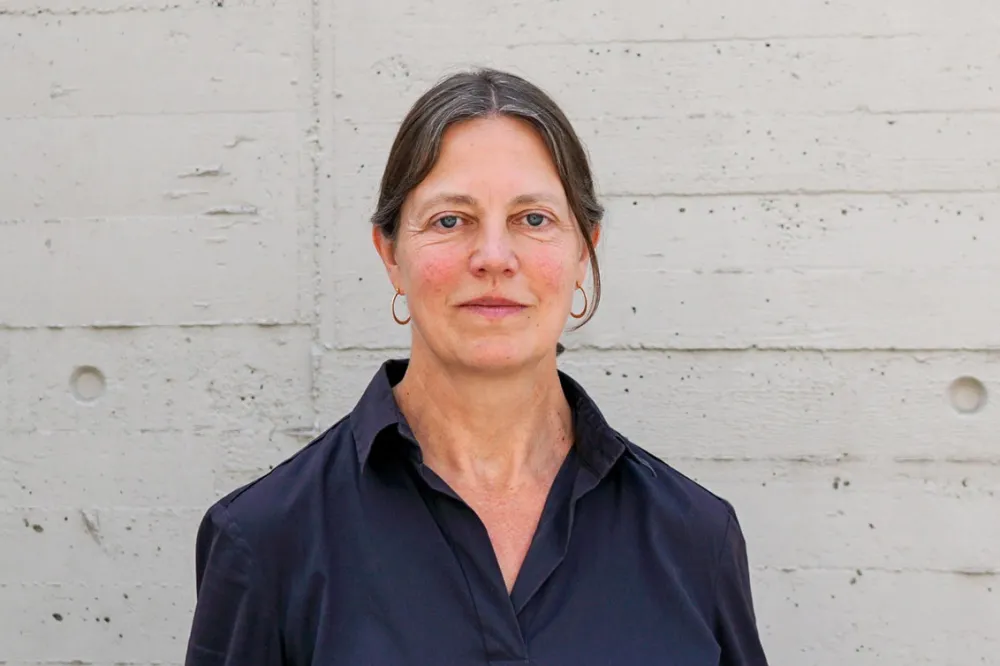 A woman with her hair tied back and wearing a dark blue blouse stands in front of a light-colored concrete wall and looks straight into the camera.