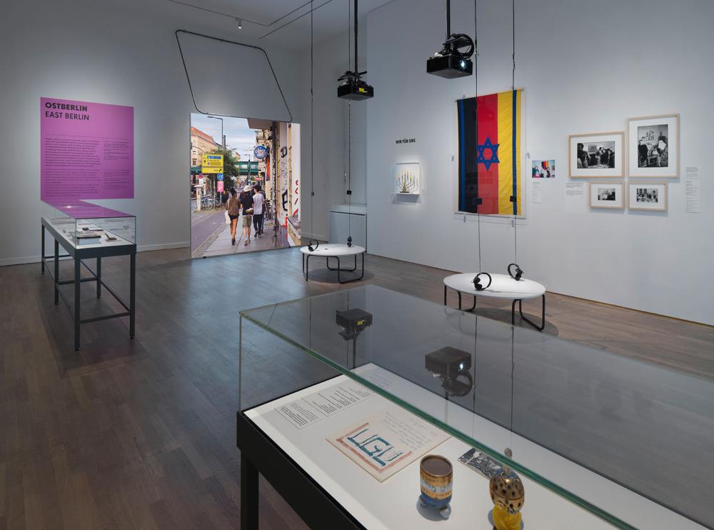 View into an exhibition room on the subject of East Berlin with showcases, paintings, canvas and headphones.