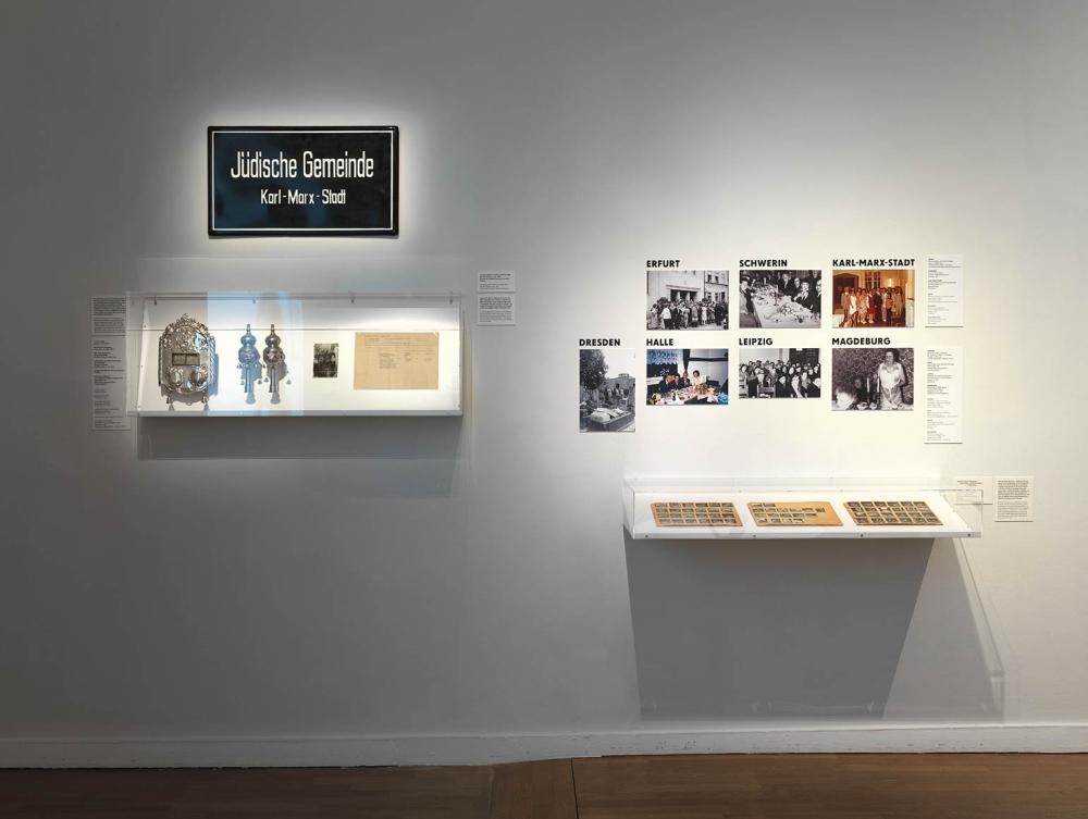 Blick an eine Ausstellungswand mit einem Schild der Jüdischen Gemeinde Karl-Marx-Stadt, Zeremonialobjekten in einer Vitrine und Fotos von weiteren jüdischen Gemeinden in der DDR.