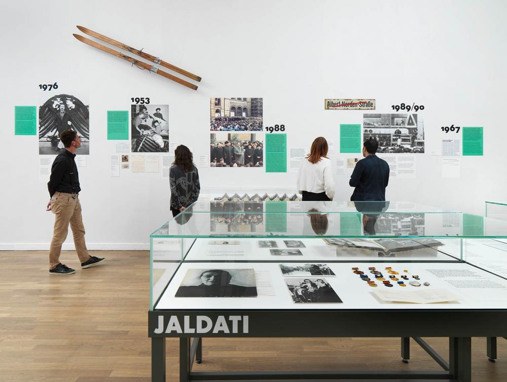 View into an exhibition room where people are looking at an illustrated timeline on the wall, above them a few wooden skis, in the foreground a display case with the inscription Jaldati.