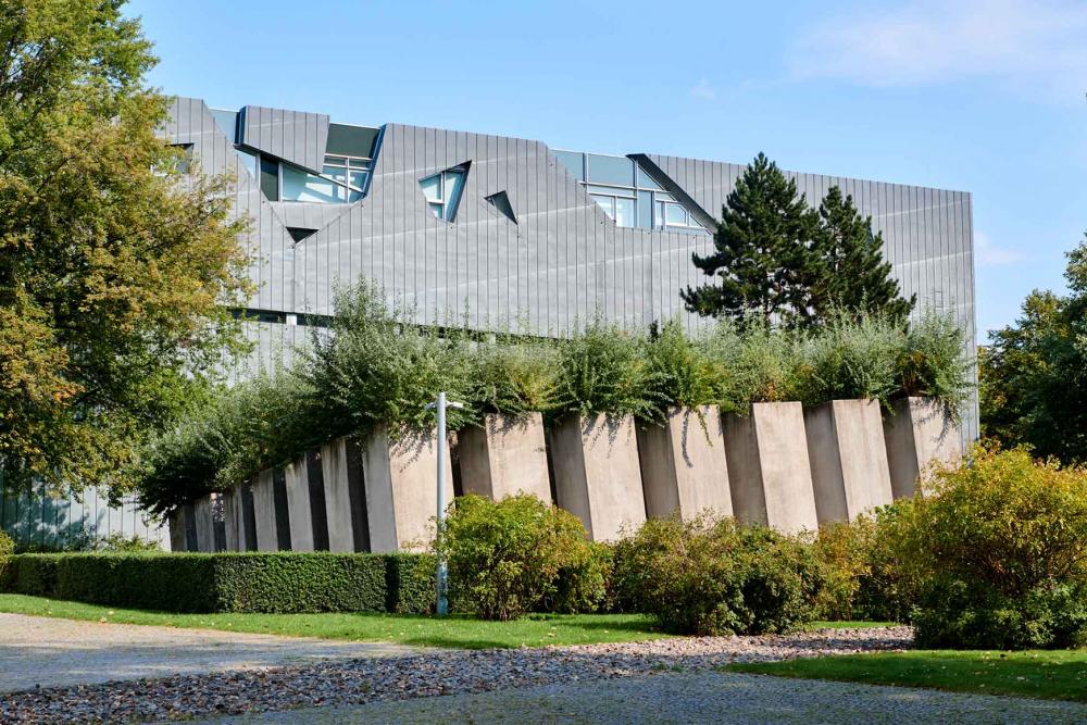 New building from metal elements, in front of it overgrown stems behind a hedge.