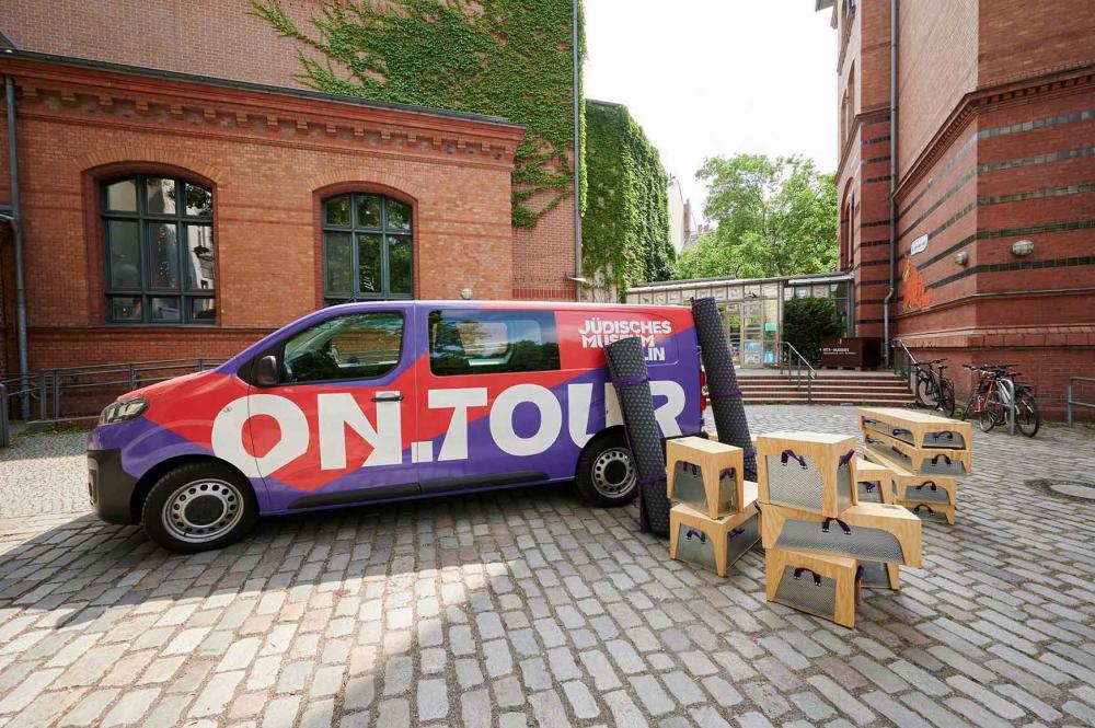 A red and purple bus on a paved yard, in front of it boxes made of wood and metal