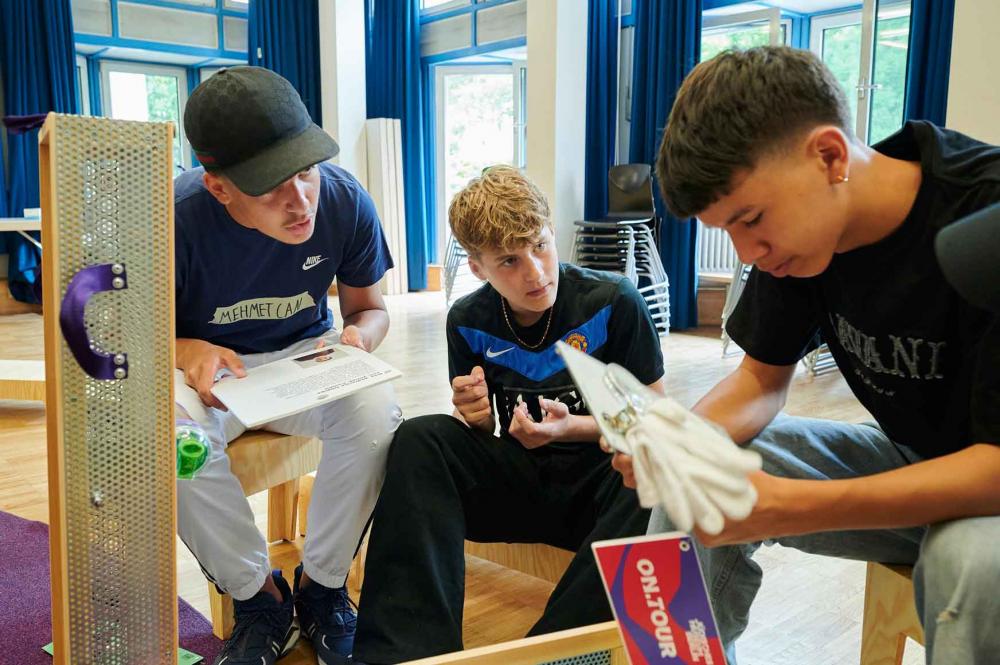 A teenager is holding a white square with a magnifying glass and white gloves. Two teenagers look at him.