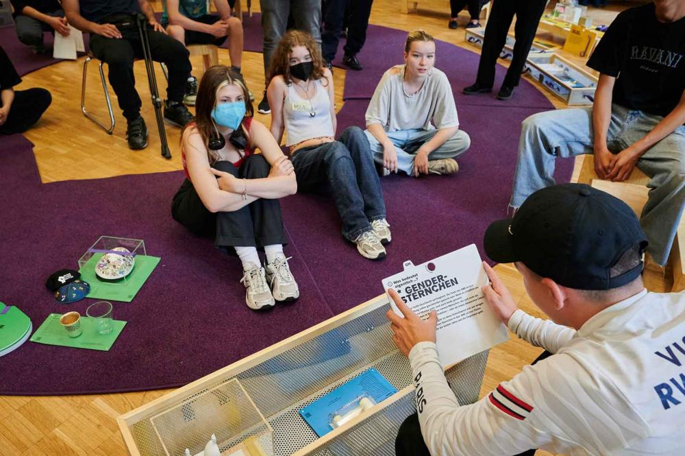 Jugendliche sitzen auf einem lila kreisförmigen Teppich. Vor ihnen sitzt ein Jugendlicher, der eine weiße Karte in der Hand trägt, die mit Gendersternchen beschriftet ist.