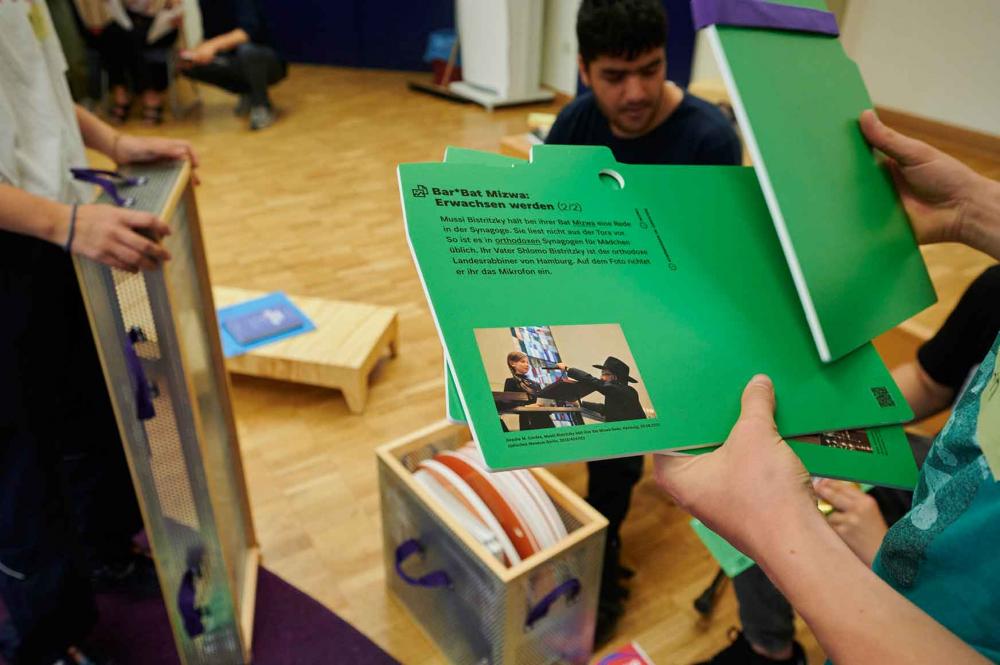 Hände halten grüne Karten in der Hand. Darauf zu sehen ist eine Fotografie und ein Text, der mit „Bar*Bat Mizwa: Erwachsen werden“ übertitelt ist.