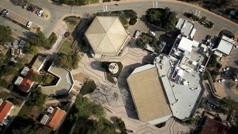 Blick aus der Vogelperspektive auf ein achteckiges Gebäude, eine größere Halle und mehrere Reihenhäuser mit roten Dächern. Sie sind umgeben von Bäumen, Vorgärten und Zufahrtswegen und Parkplätzen.