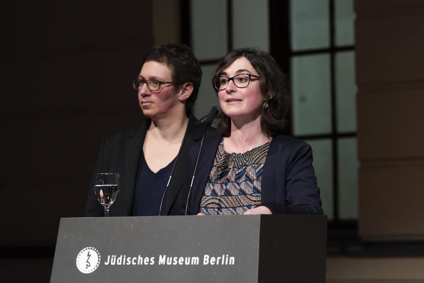 Alina Gromova steht an einem Pult auf dem das Logo und der Schriftzug des Jüdischen Museums stehen. Auf dem Pult steht ein Wasserglas. Hinter ihr steht Susan Kamel.