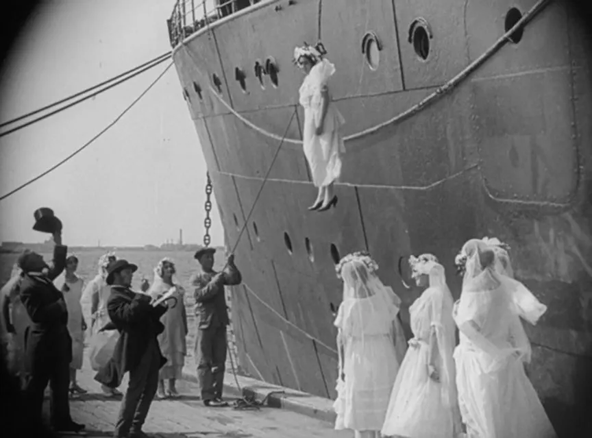 Auf einem schwarz-weißen Filmstill ist eine Menschenmenge vor einem Schiff zu sehen. Links stehen Männer im Anzug, rechts stehen drei Frauen in weißen Kleidern. Alle blicken auf zu einer Frau in einem Brautkleid, die an einem Seil vor dem Schiff hängt.