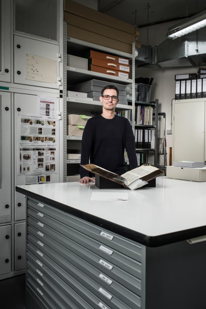 Ein Mann steht hinter einem Arbeitstisch in einem Atelier; vor ihm liegt ein altes aufgeschlagenes Buch