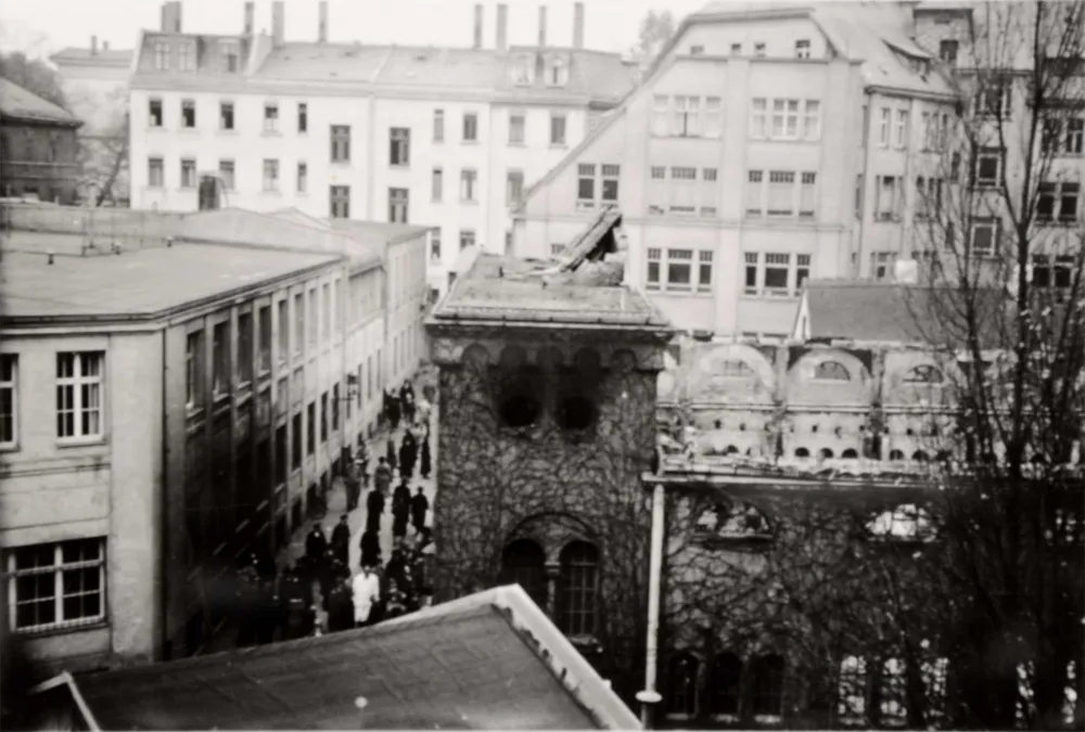 Das Fotot zeigt eine zerstörte Synagoge, deren Dach fehlt, es scheint verbrannt zu sein, Trümmer liegen auf dem noch bestehenden Eingangsportal, in der Staße davor befinden sich viele Passanten.