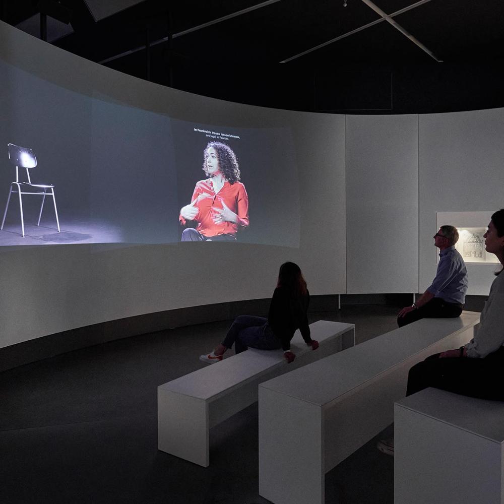 People on benches in front of a round screen on the wall, on it an empty chair and a chair with a female rabbi sitting on it