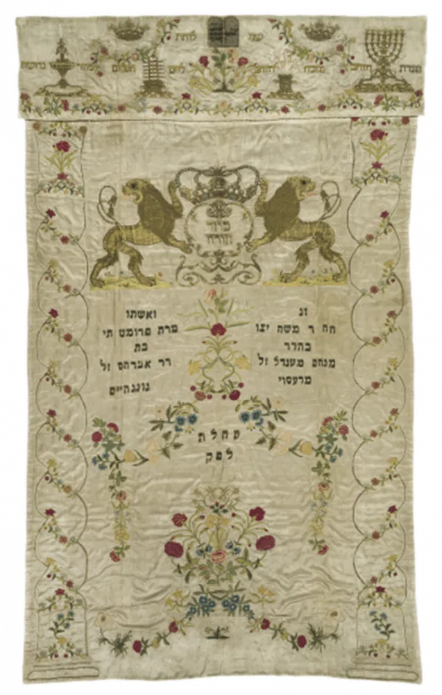 Embroidered white Torah curtain with numerous decorations and two lions and a crown over the Hebrew dedication inscription.