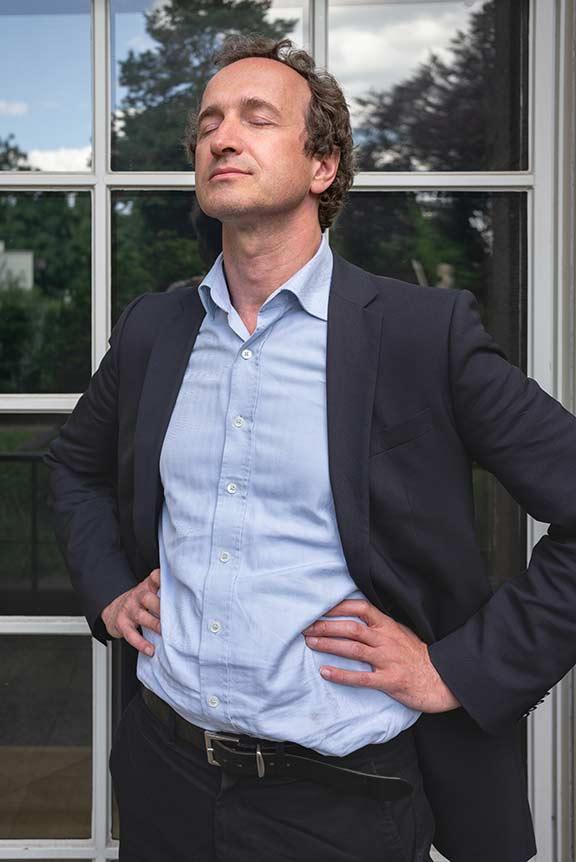 A man in a light blue shirt and dark blue suit stands in half-profile in front of a window pane in which trees are reflected, his eyes are closed, his hands are at his sides, a smile plays around his mouth.